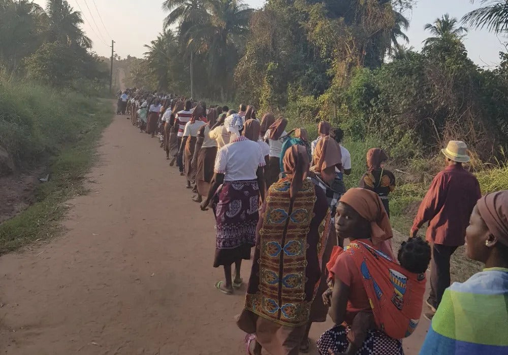 A line of African people standing along a dirt road surrounded by foliage