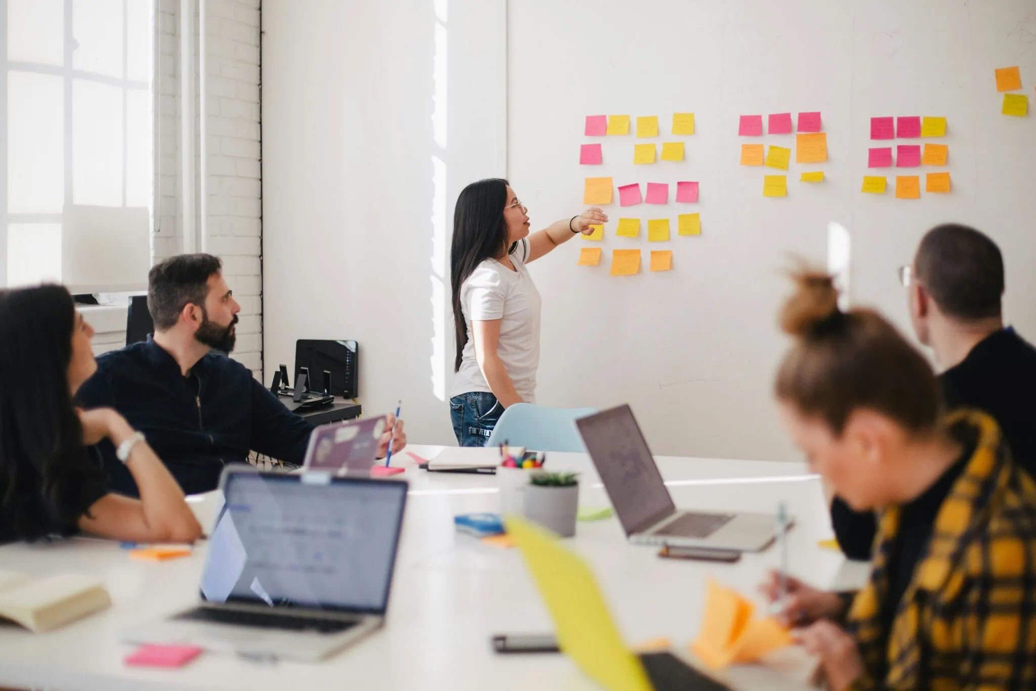A group of people in a meeting while one person points at orange and yellow sticky notes on the wall